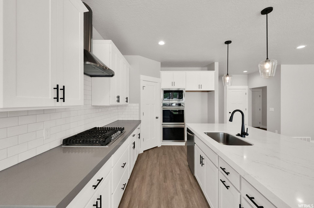 Kitchen featuring hardwood floors, gas cooktop, fume extractor, double oven, dishwasher, kitchen island sink, white cabinetry, pendant lighting, and light countertops