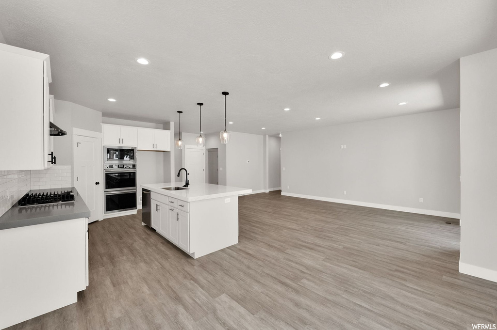 Kitchen with dishwasher, microwave, double oven, pendant lighting, light parquet floors, white cabinetry, a kitchen island with sink, and light countertops