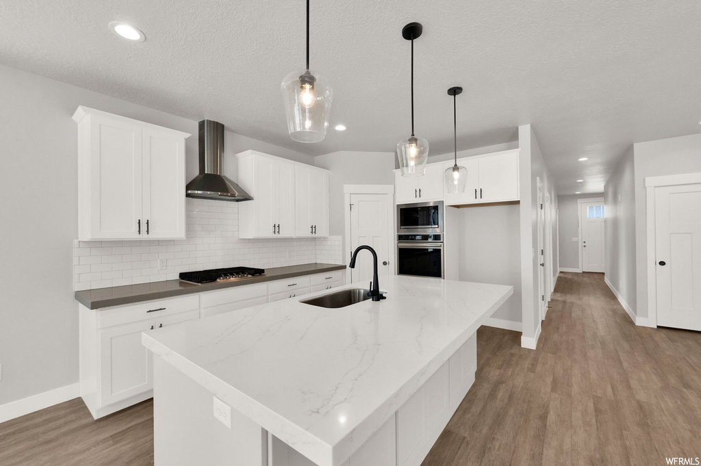 Kitchen featuring wood-type flooring, oven, extractor fan, a kitchen island with sink, white cabinets, pendant lighting, and light countertops