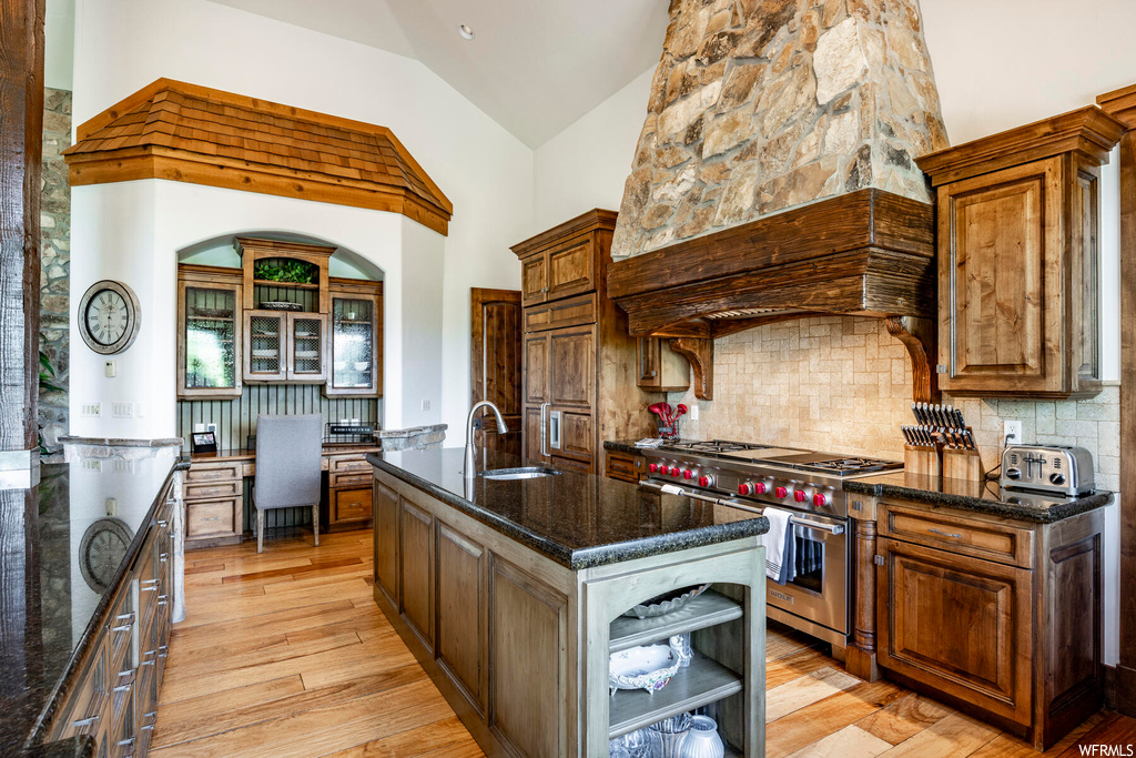 Kitchen featuring a kitchen island, vaulted ceiling, gas range oven, stainless steel finishes, light hardwood flooring, dark stone countertops, and brown cabinets