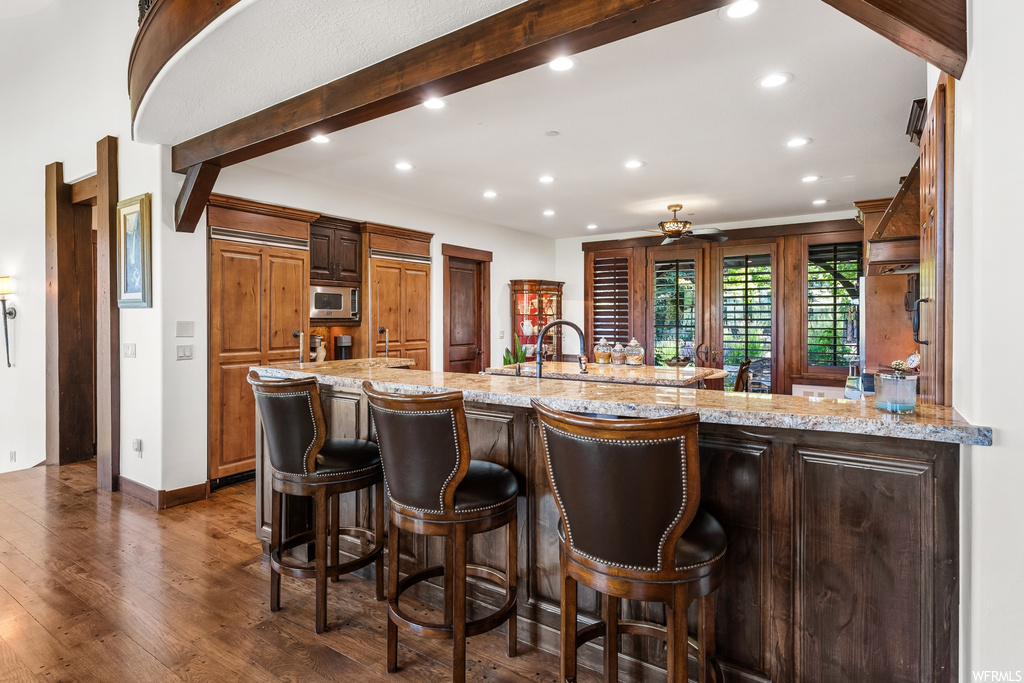 Kitchen featuring a ceiling fan, hardwood floors, a kitchen bar, natural light, microwave, kitchen island sink, and light stone countertops