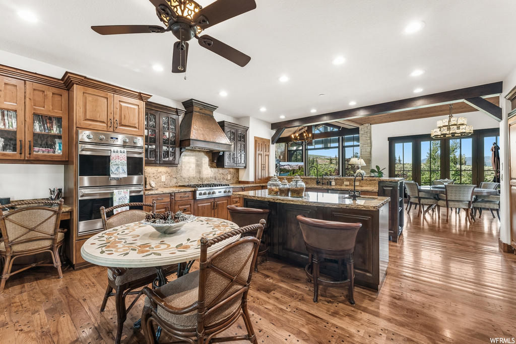 Kitchen with natural light, a notable chandelier, a kitchen breakfast bar, gas cooktop, range hood, stainless steel double oven, light hardwood floors, and kitchen island sink