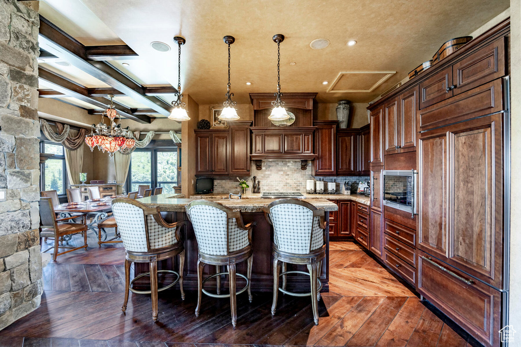 Kitchen featuring light stone counters, coffered ceiling, built in appliances, hanging light fixtures, and a center island with sink