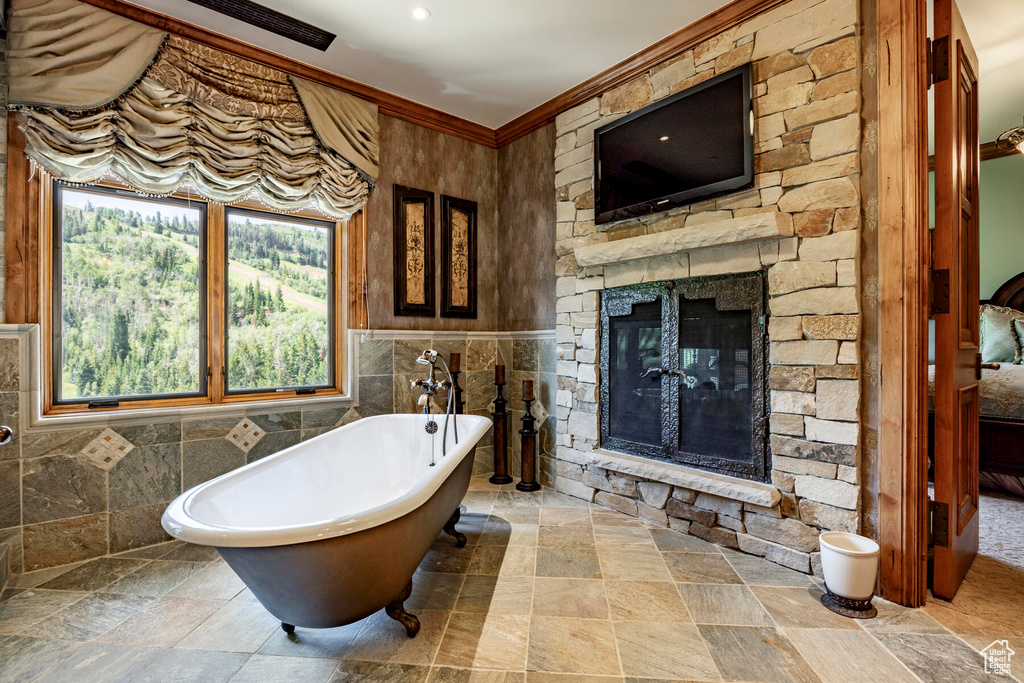 Bathroom with a tub, ornamental molding, tile walls, a stone fireplace, and tile floors