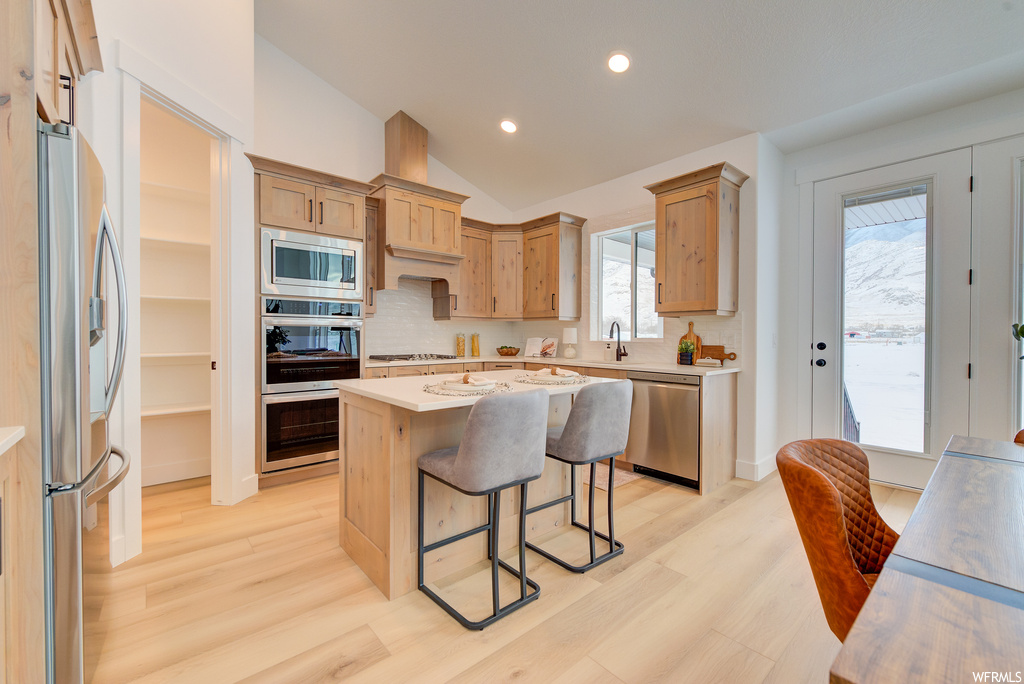 Kitchen featuring vaulted ceiling, light hardwood / wood-style floors, appliances with stainless steel finishes, a center island, and tasteful backsplash