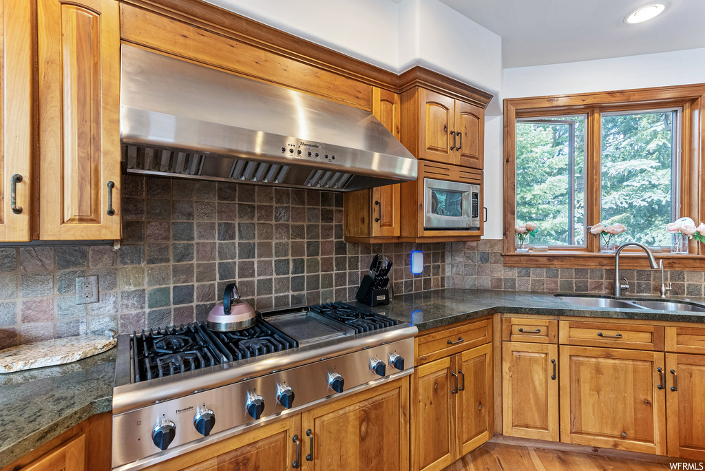Kitchen featuring backsplash, dark countertops, hardwood flooring, stainless steel gas stovetop, brown cabinets, and wall chimney range hood