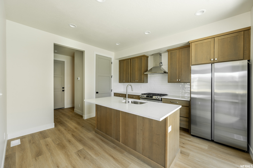 Kitchen with high end refrigerator, light hardwood floors, light countertops, wall chimney range hood, and backsplash
