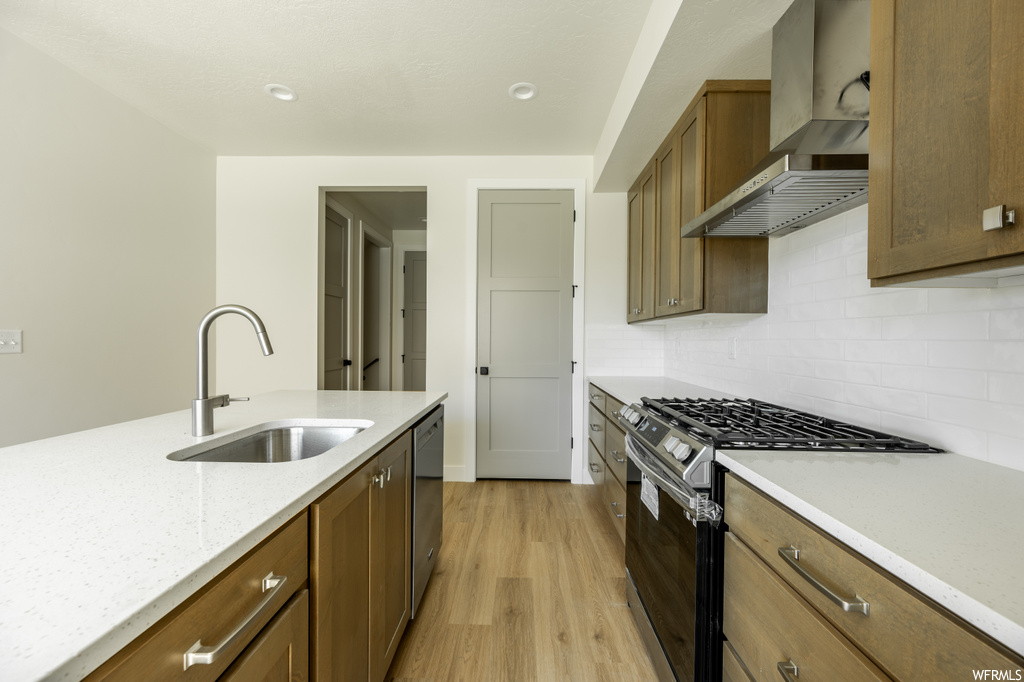 Kitchen with light countertops, wall chimney exhaust hood, backsplash, light hardwood flooring, and stainless steel appliances