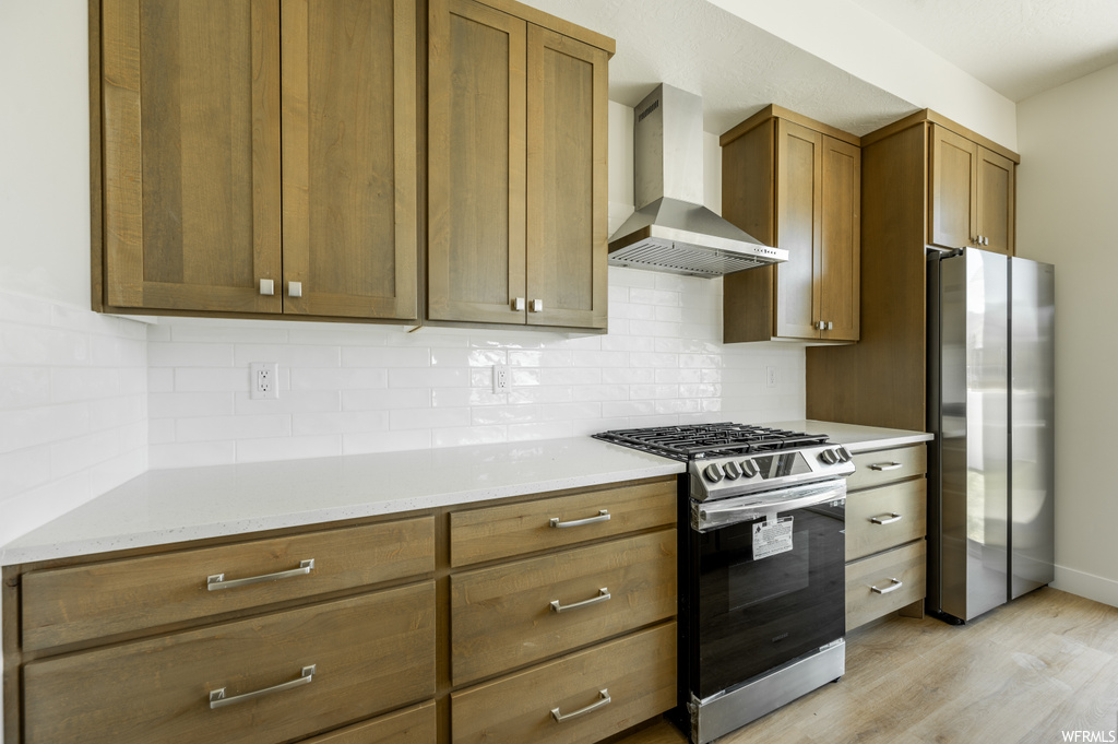 Kitchen with backsplash, light countertops, brown cabinets, wall chimney range hood, light hardwood floors, and stainless steel appliances