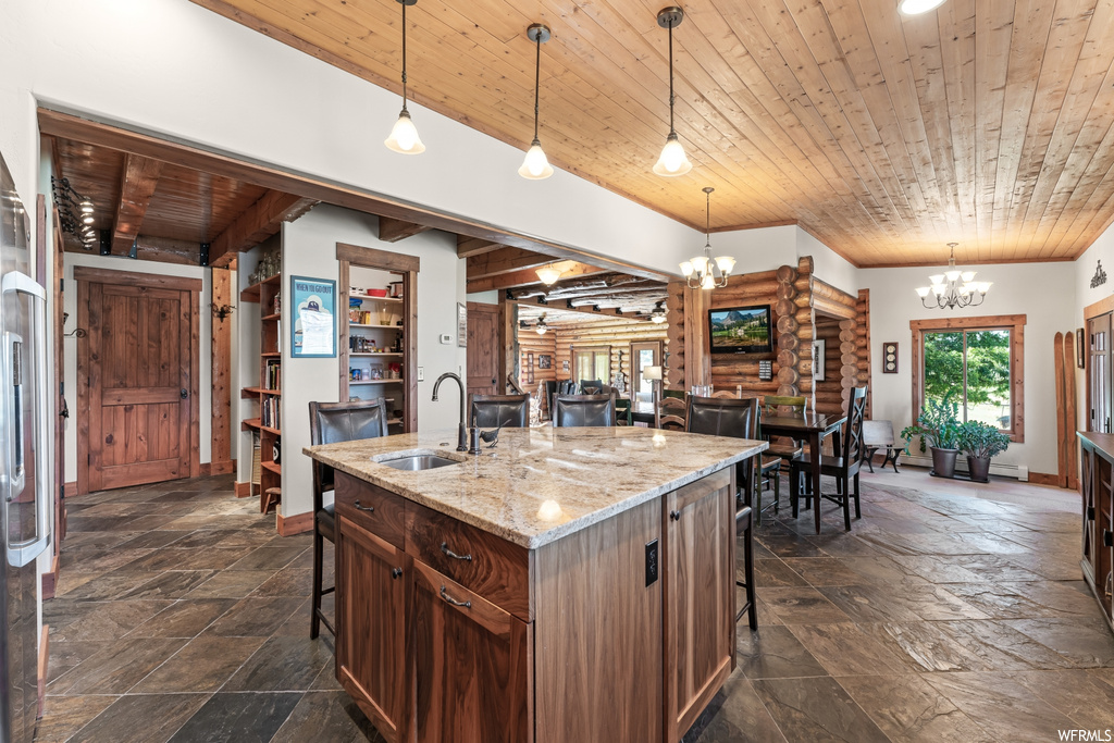 Kitchen with pendant lighting, a chandelier, log walls, dark tile flooring, light stone countertops, wood ceiling, and kitchen island with sink