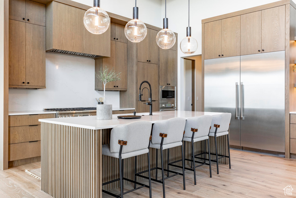 Kitchen featuring built in appliances, a kitchen bar, light hardwood / wood-style flooring, a kitchen island with sink, and pendant lighting