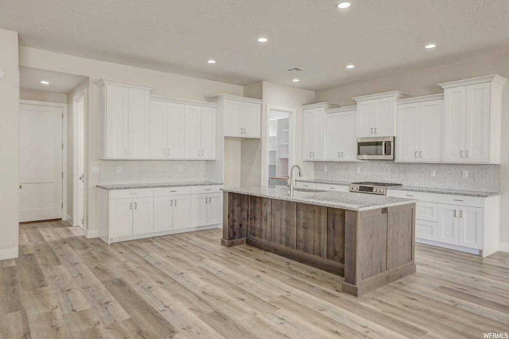 Kitchen featuring white cabinets, a kitchen island with sink, light granite-like countertops, backsplash, and light hardwood floors