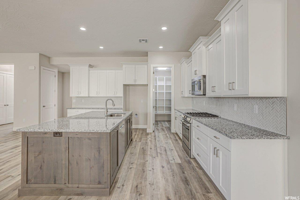 Kitchen with white cabinets, light parquet floors, a kitchen island with sink, backsplash, light stone countertops, and stainless steel appliances