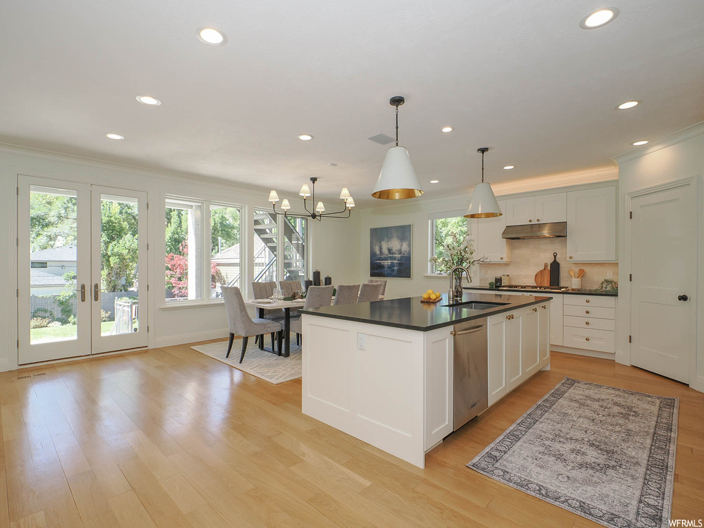 Kitchen featuring crown molding, french doors, backsplash, dark countertops, white cabinets, light hardwood flooring, decorative light fixtures, a kitchen island with sink, and stainless steel dishwasher