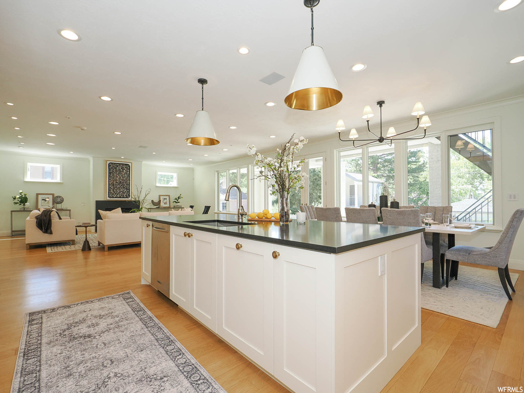 Kitchen featuring pendant lighting, dark granite-like countertops, kitchen island with sink, stainless steel dishwasher, and light hardwood floors