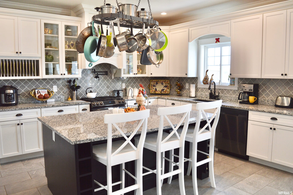 Kitchen featuring stone counters, an island with sink, light tile floors, black dishwasher, a kitchen island, high end range oven, backsplash, crown molding, and white cabinetry