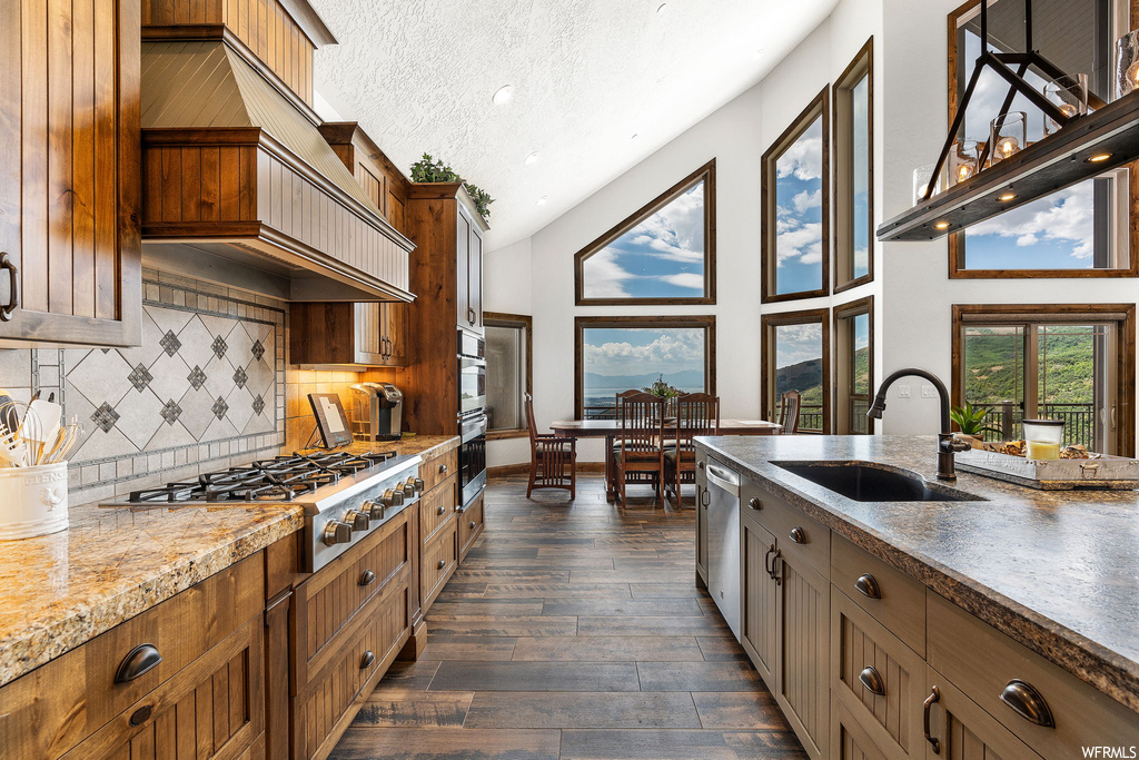 Kitchen featuring light stone counters, brown cabinets, lofted ceiling, a textured ceiling, appliances with stainless steel finishes, wood-type flooring, backsplash, and a high ceiling