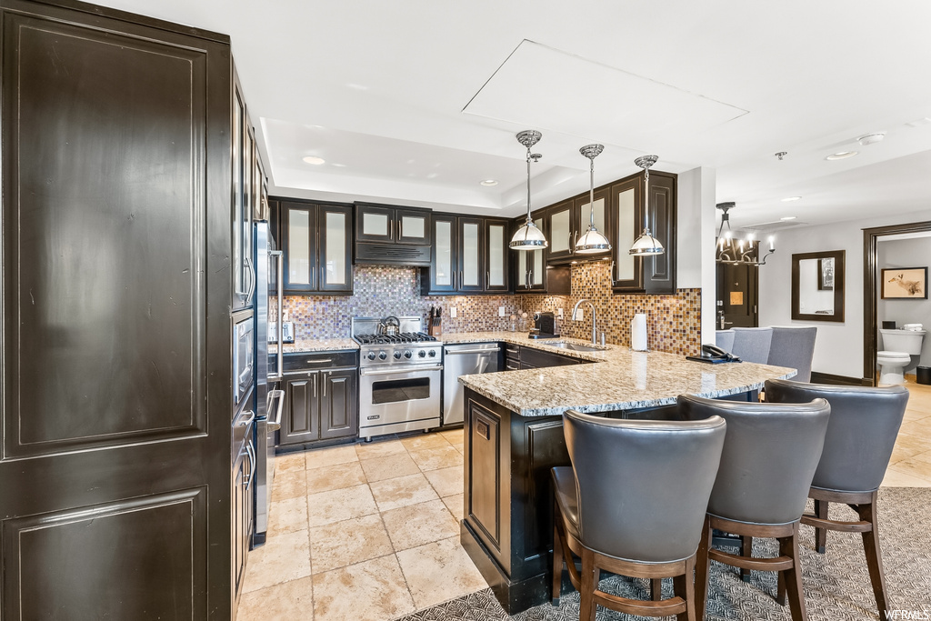 Kitchen with dark brown cabinets, premium range, hanging light fixtures, backsplash, light stone countertops, and light tile flooring