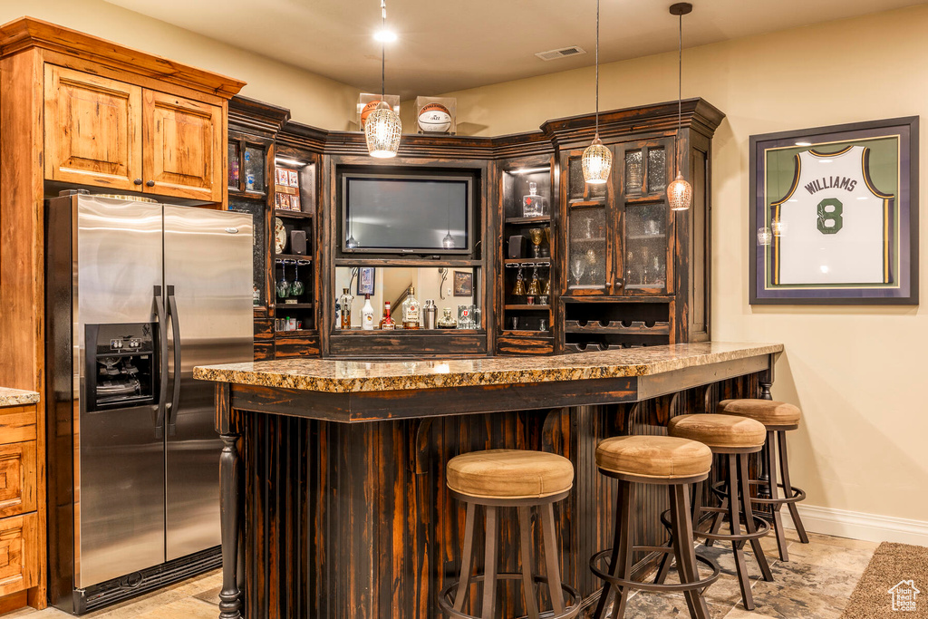 Bar with dark stone counters, stainless steel fridge with ice dispenser, light tile flooring, and decorative light fixtures