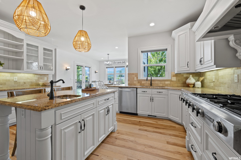 Kitchen featuring light parquet floors, backsplash, plenty of natural light, white cabinets, stone counters, custom exhaust hood, decorative light fixtures, and appliances with stainless steel finishes
