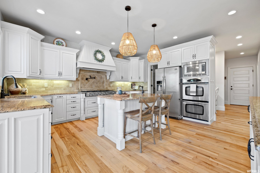 Kitchen with white cabinets, light hardwood flooring, appliances with stainless steel finishes, light stone countertops, decorative light fixtures, backsplash, custom exhaust hood, and a center island