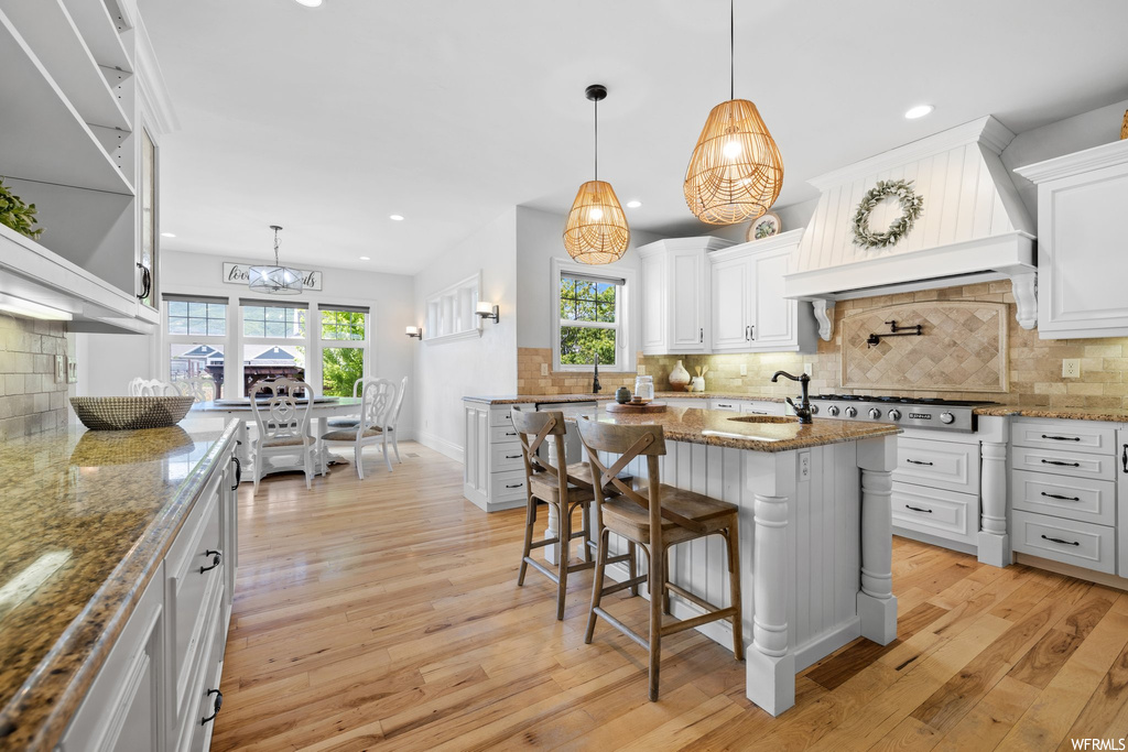 Kitchen with a kitchen island, dark stone counters, premium range hood, backsplash, stainless steel gas cooktop, hanging light fixtures, light hardwood flooring, a healthy amount of sunlight, and white cabinetry