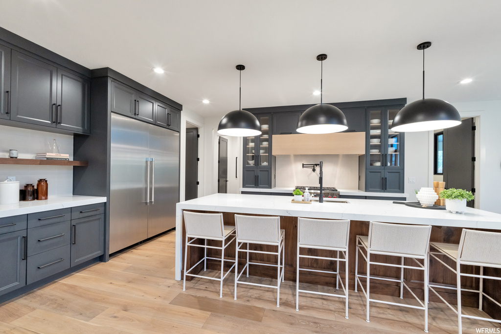 Kitchen featuring light hardwood flooring, decorative light fixtures, stainless steel built in fridge, dark brown cabinetry, light countertops, and a center island