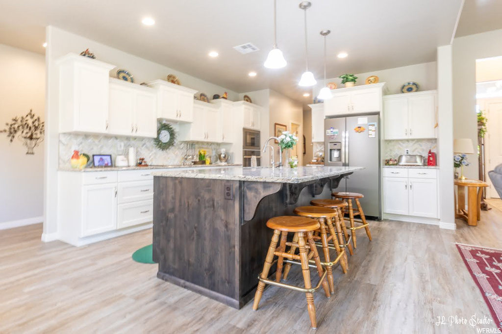Kitchen featuring stainless steel refrigerator with ice dispenser, backsplash, light countertops, hanging light fixtures, light hardwood flooring, white cabinetry, and a center island