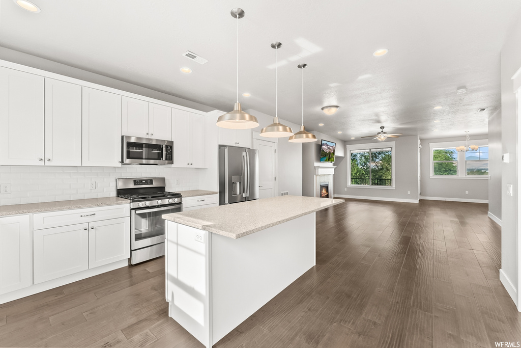 Kitchen featuring stainless steel appliances, backsplash, an island with sink, white cabinets, ceiling fan, wood-type flooring, and hanging light fixtures