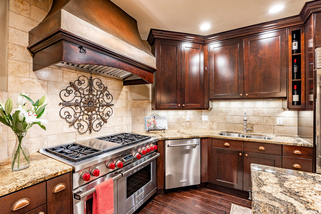 Kitchen with hardwood floors, stainless steel appliances, dark brown cabinets, premium range hood, backsplash, and light stone counters