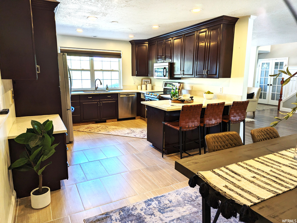 Kitchen featuring dark brown cabinets, light countertops, appliances with stainless steel finishes, light tile floors, and a center island