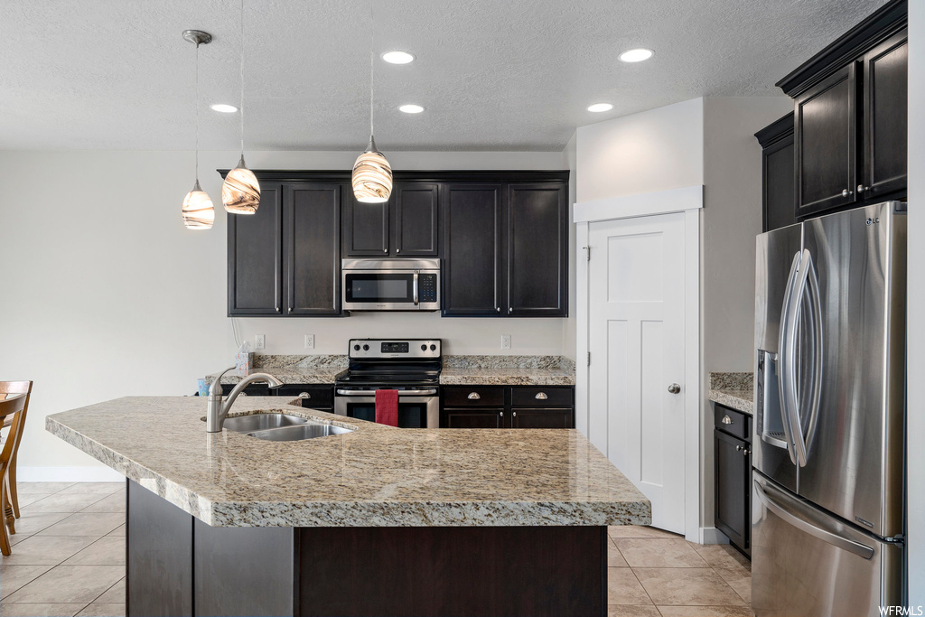 Kitchen featuring a textured ceiling, light tile floors, dark brown cabinetry, stone counters, pendant lighting, and appliances with stainless steel finishes