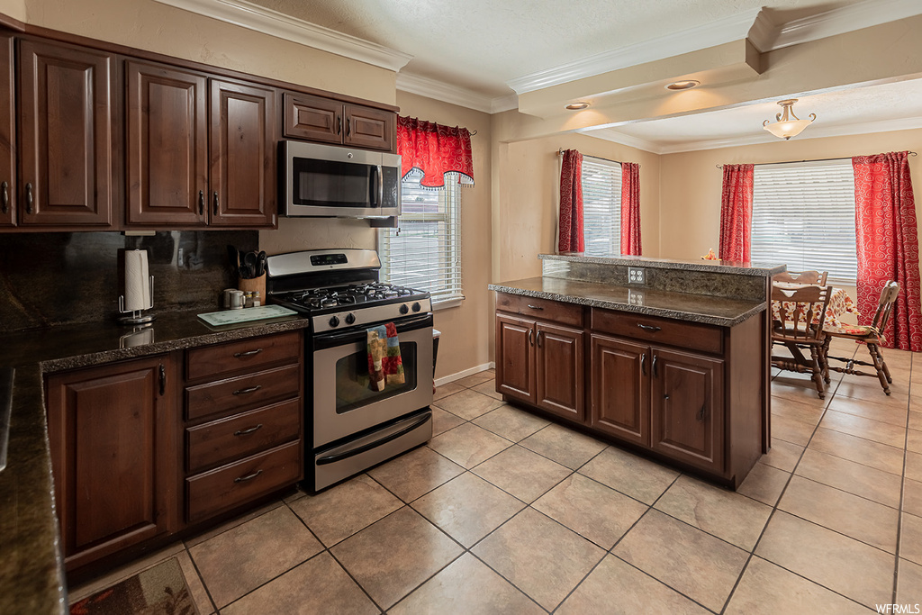 Kitchen featuring appliances with stainless steel finishes, backsplash, light tile flooring, dark stone counters, and ornamental molding