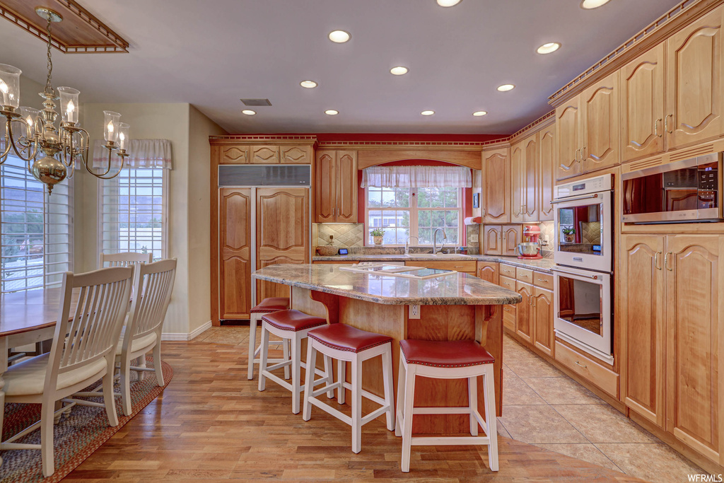 Kitchen featuring light granite-like countertops, a kitchen island, backsplash, brown cabinets, a chandelier, light hardwood flooring, and white double oven