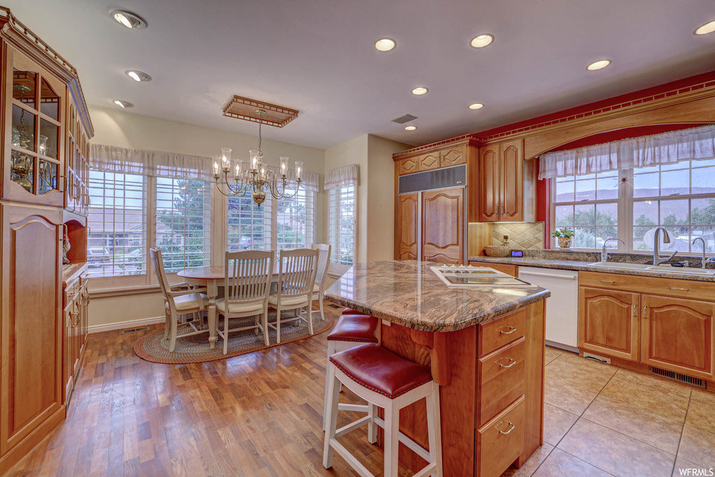 Kitchen with brown cabinets, a notable chandelier, a center island with sink, stone countertops, a kitchen island, a wealth of natural light, white dishwasher, backsplash, and light hardwood floors