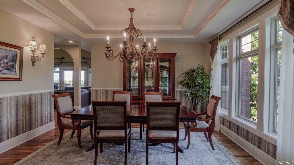Wood floored dining room with plenty of natural light, a raised ceiling, a notable chandelier, and crown molding