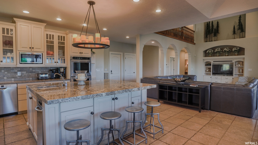 Kitchen featuring kitchen island with sink, backsplash, appliances with stainless steel finishes, light tile floors, a center island, and dark granite-like countertops