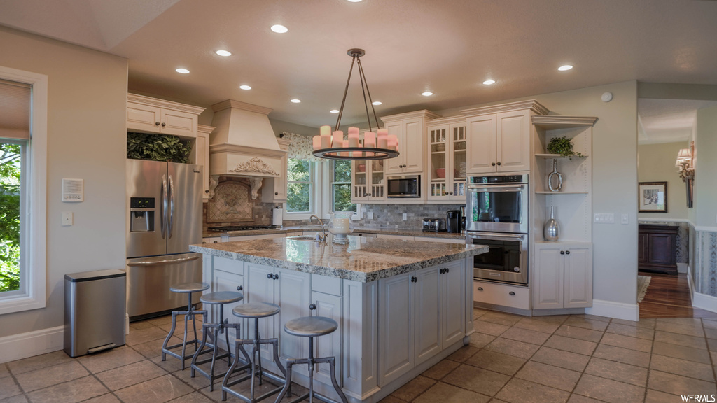 Kitchen featuring light tile flooring, kitchen island with sink, plenty of natural light, backsplash, appliances with stainless steel finishes, light stone countertops, custom exhaust hood, white cabinetry, and a center island