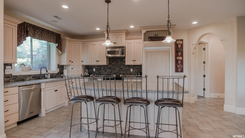 Kitchen with stainless steel appliances, a kitchen island, backsplash, light tile floors, white cabinetry, pendant lighting, and dark granite-like countertops