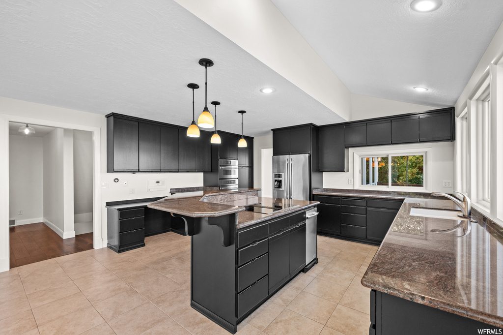 Kitchen featuring an island with sink, high end fridge, dark brown cabinets, vaulted ceiling, hanging light fixtures, light countertops, and light tile flooring