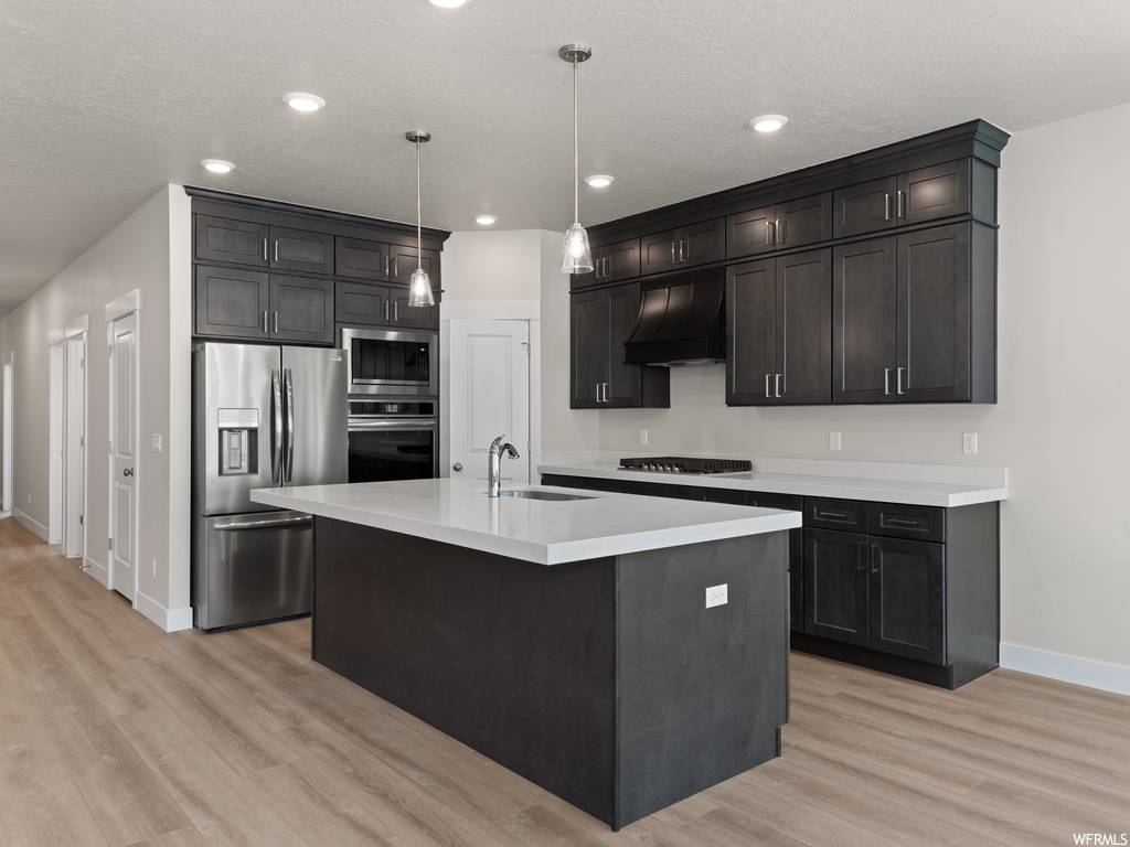Kitchen featuring light hardwood flooring, dark brown cabinets, appliances with stainless steel finishes, a kitchen island with sink, decorative light fixtures, custom range hood, and light countertops