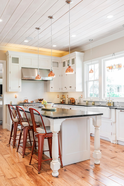 Kitchen with backsplash, white cabinetry, a center island with sink, hanging light fixtures, wall chimney range hood, dark stone countertops, a center island, light hardwood floors, and crown molding