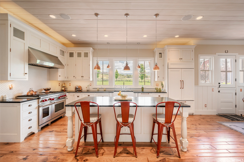 Kitchen with backsplash, dark countertops, high end appliances, a center island, light hardwood floors, pendant lighting, white cabinets, and crown molding