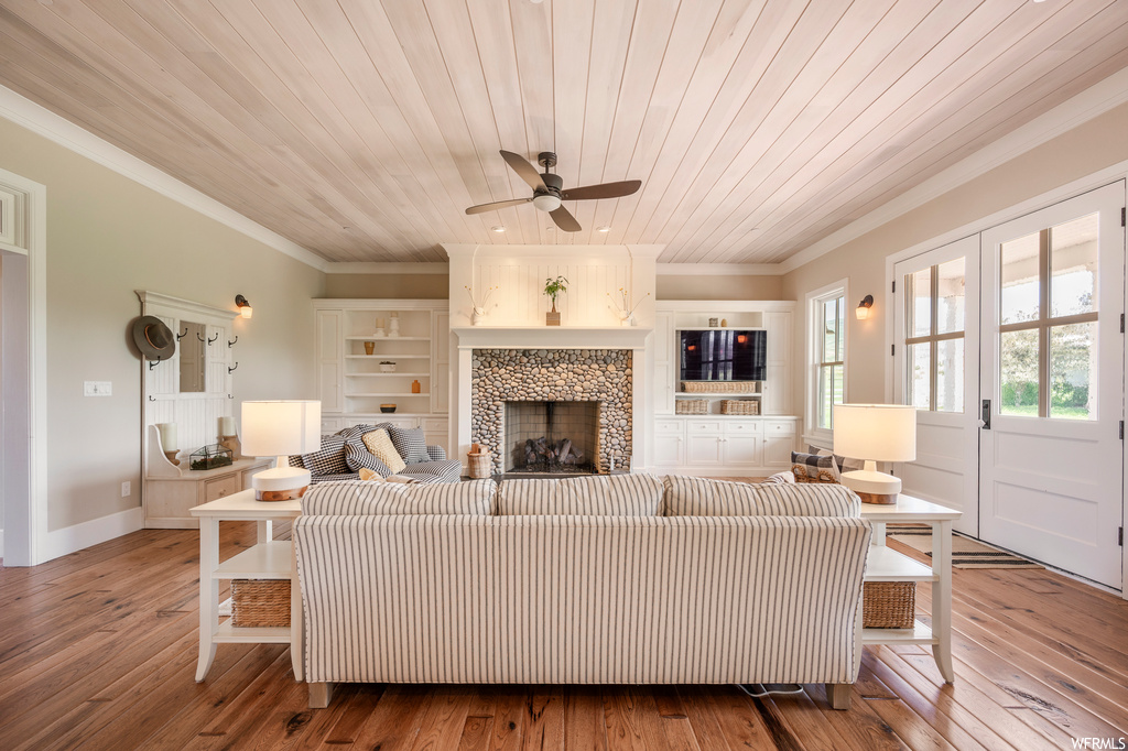 Hardwood floored living room featuring ceiling fan, built in features, wood ceiling, ornamental molding, and a fireplace
