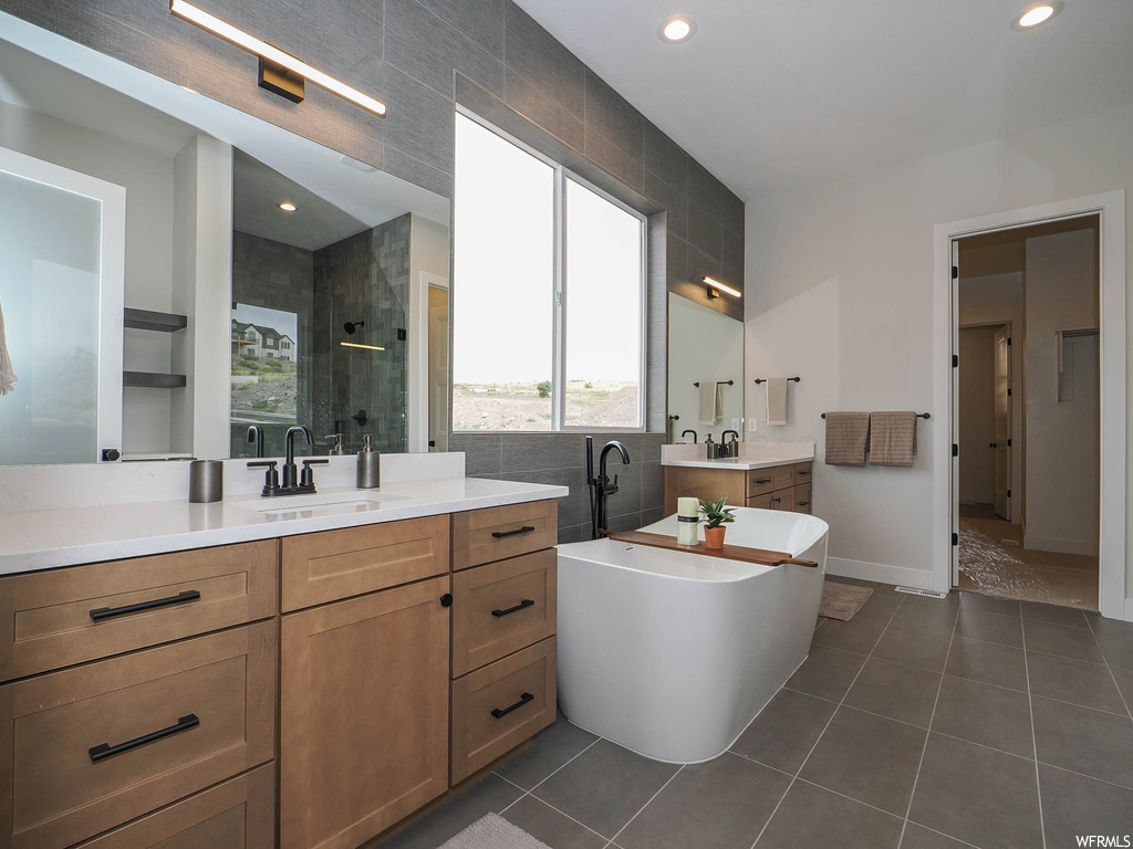 Bathroom featuring dual bowl vanity, mirror, dark tile flooring, separate shower and tub enclosures, and tile walls