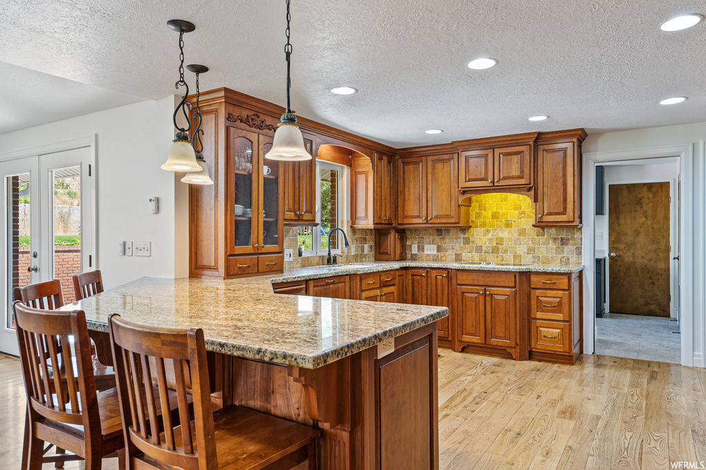 Kitchen with brown cabinets, light stone counters, pendant lighting, backsplash, a textured ceiling, plenty of natural light, light hardwood floors, and a center island
