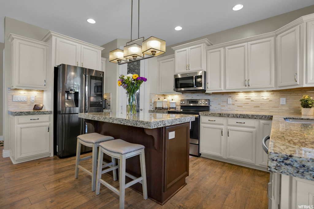 Kitchen with stainless steel appliances, light stone counters, pendant lighting, backsplash, white cabinetry, hardwood flooring, and a center island