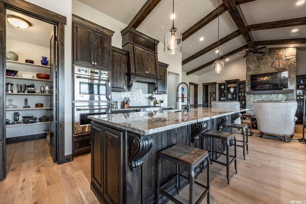 Kitchen featuring light hardwood flooring, light stone counters, dark brown cabinetry, stainless steel double oven, vaulted ceiling with beams, hanging light fixtures, backsplash, and a kitchen island