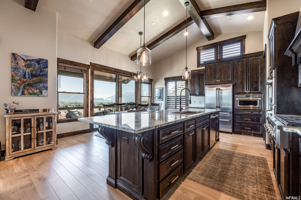 Kitchen with a high ceiling, light hardwood flooring, granite-like countertops, decorative light fixtures, dark brown cabinets, lofted ceiling with beams, stainless steel oven, and kitchen island with sink