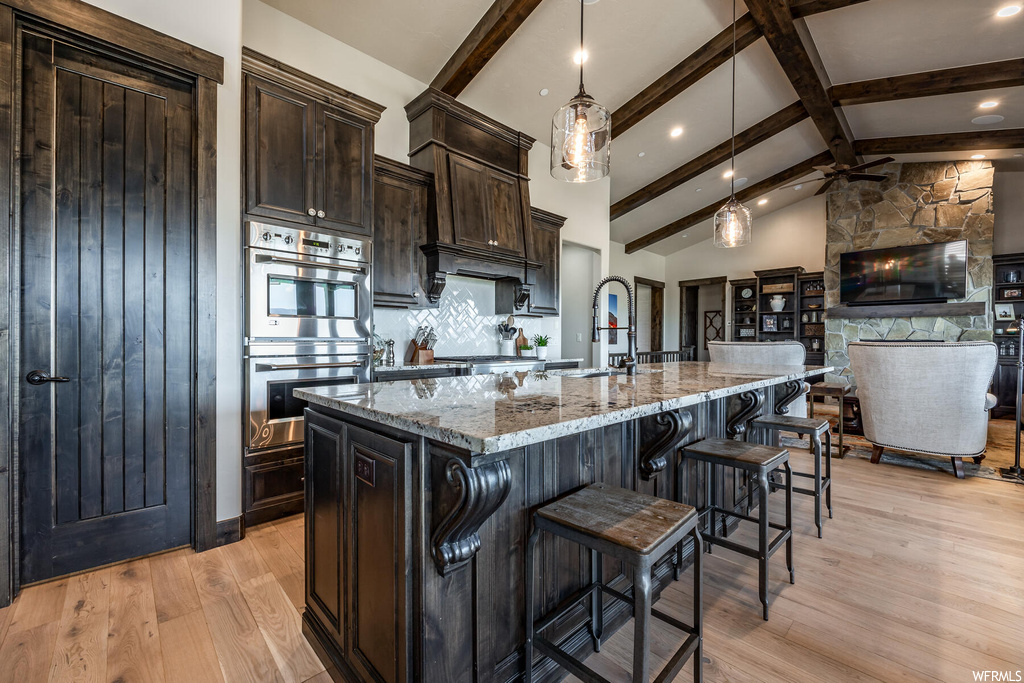 Kitchen with granite-like countertops, dark brown cabinets, light hardwood flooring, backsplash, vaulted ceiling with beams, stainless steel double oven, pendant lighting, kitchen island with sink, and a kitchen island